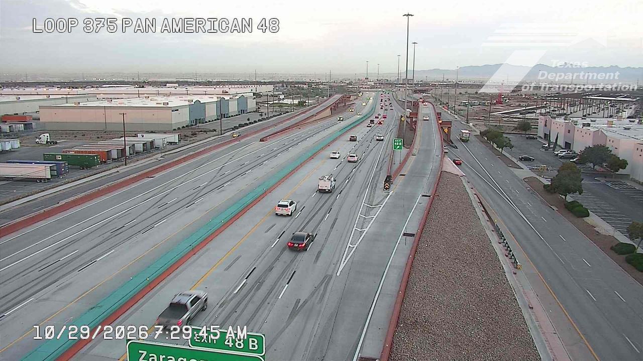 Light traffic flows along a multi-lane highway with commercial buildings and freight trucks on the adjacent ramps; no visible incidents or road work.