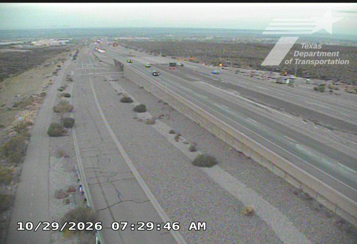 A multi-lane highway with light traffic flows under overcast skies, bordered by desert vegetation and a concrete barrier.
