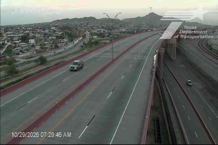 A multi-lane highway with light traffic, featuring a single vehicle moving in the left lane and another on the adjacent lane, with distant hills and urban development visible under clear skies.