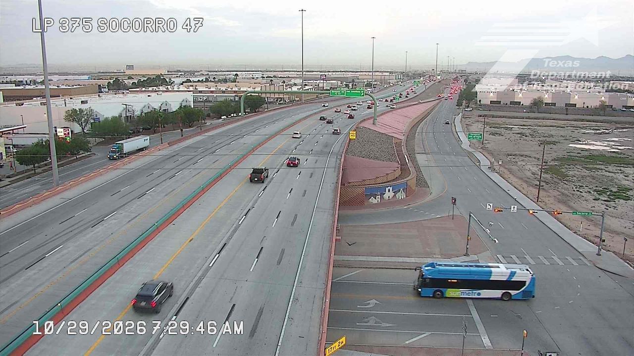 Multi-lane highway with moderate traffic flow, featuring a blue and white bus turning at an intersection and a semi-truck on the adjacent lane; overcast skies and distant industrial buildings.