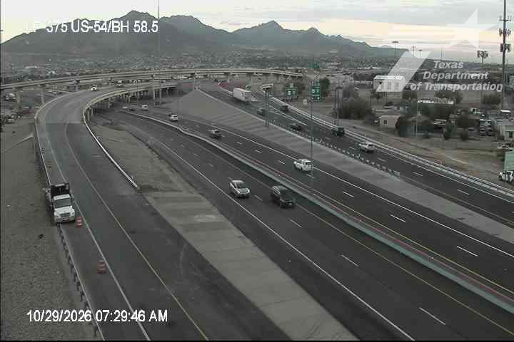 Multi-lane highway with moderate traffic flow under overpasses, with a semi truck parked on the shoulder near construction cones; distant mountains under overcast skies.