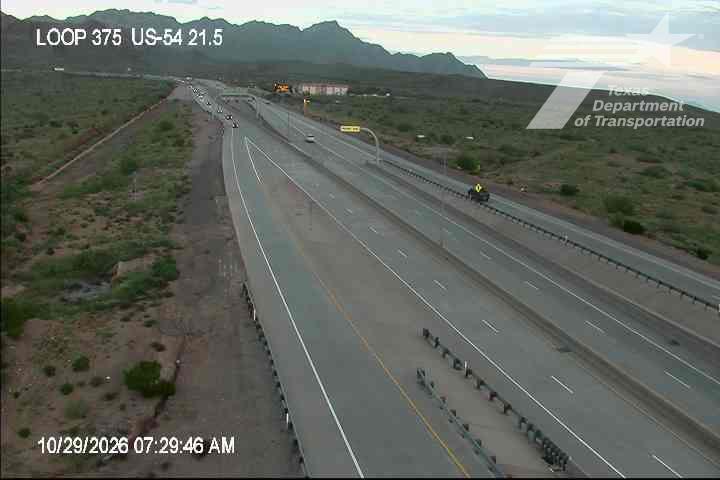 A multi-lane highway stretches through a desert landscape with distant mountains, showing light traffic with a few vehicles moving freely; no visible work zones or incidents.
