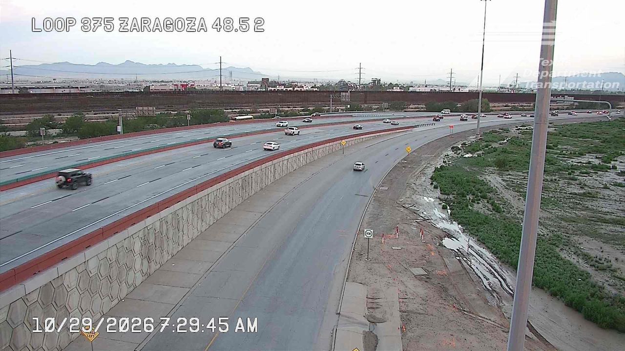 Multi-lane highway with moderate traffic flowing in both directions, separated by a concrete barrier; construction activity visible on the right side with cones and a work zone sign, under overcast skies.