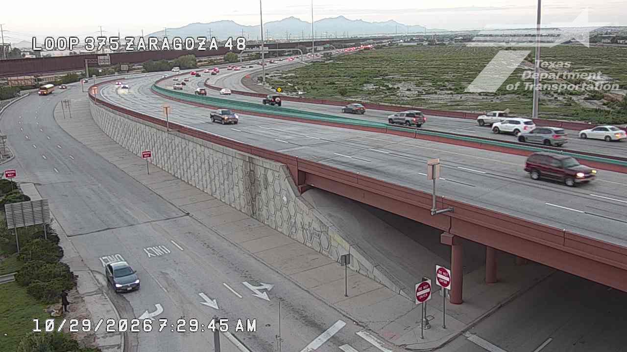 Multi-lane highway with moderate traffic flowing under an overpass; vehicles include sedans, SUVs, and a bus; weather appears clear with distant mountains visible.
