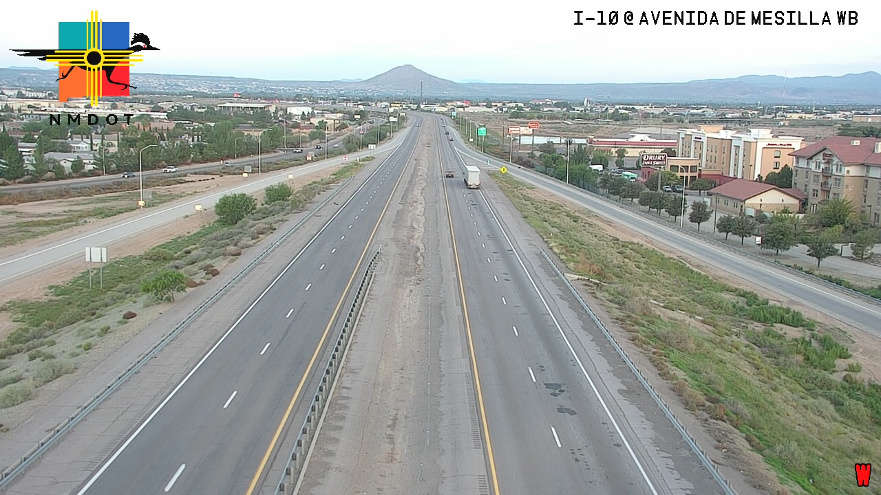 Highway I-10 at Avenida de Mesilla WB shows light traffic with a single white semi-truck traveling in the right lane, surrounded by dry grass and distant mountains under overcast skies.