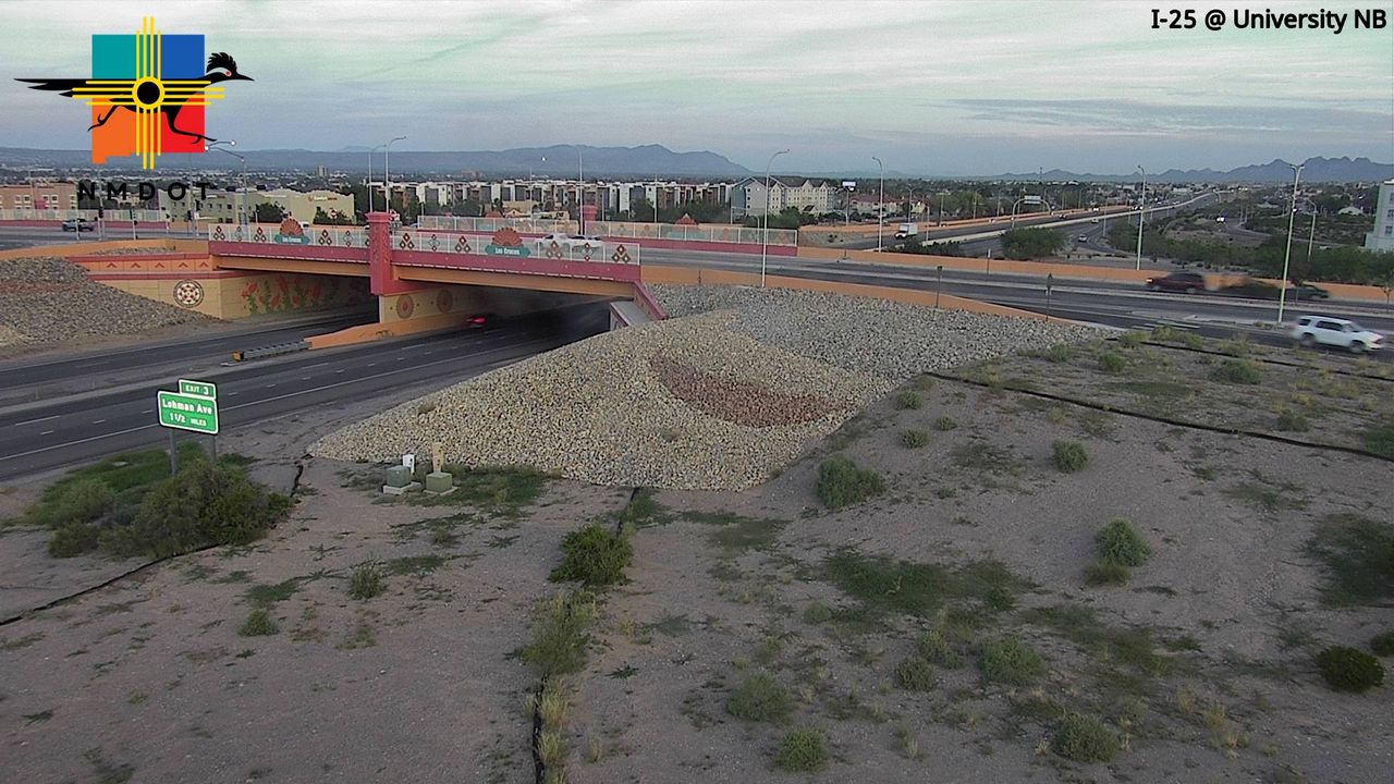 A wide highway intersection under an overpass with light traffic moving freely, set against a backdrop of distant mountains and sparse desert vegetation under an overcast sky.