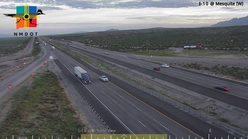 Multi-lane highway with moderate traffic flow under clear skies, featuring semi trucks and passenger vehicles moving steadily across desert terrain with distant mountains.