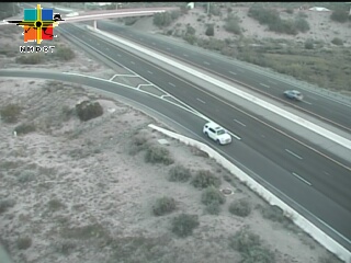 A multi-lane highway with light traffic, featuring a white SUV and a distant blue vehicle, bordered by arid desert terrain with sparse vegetation.