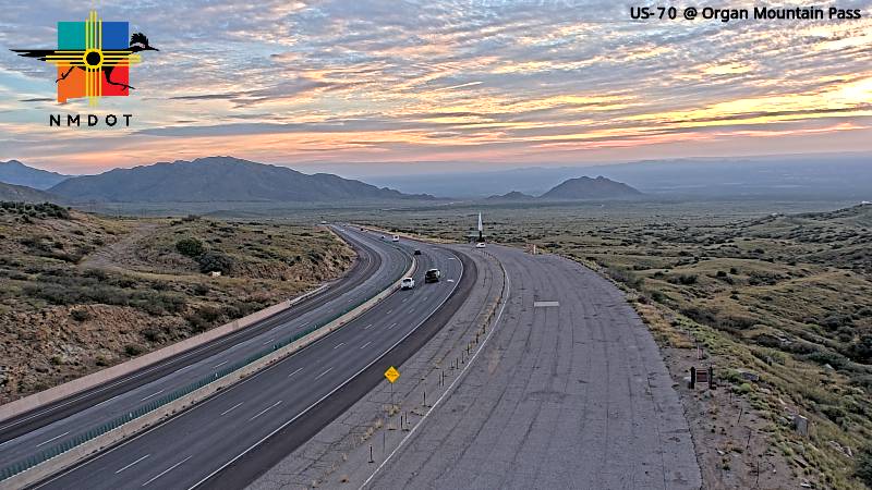 A wide highway curves through a desert landscape with distant mountains under a colorful sunset sky, with light traffic moving in both directions.