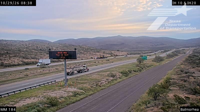 A multi-lane highway stretches through a dry, mountainous landscape with light traffic and a large truck convoy moving slowly; a digital sign warns drivers not to text while driving.