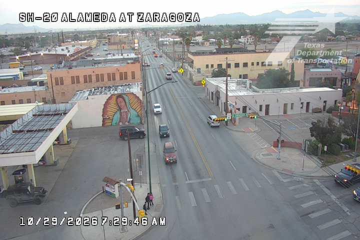 Light traffic flows through an urban intersection with a large mural on a building wall, and a few vehicles including a red SUV and a black SUV parked near a gas station.