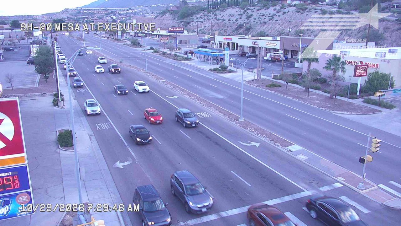 Light traffic flows through a multi-lane urban intersection with commercial buildings and a gas station on the left, under clear skies.