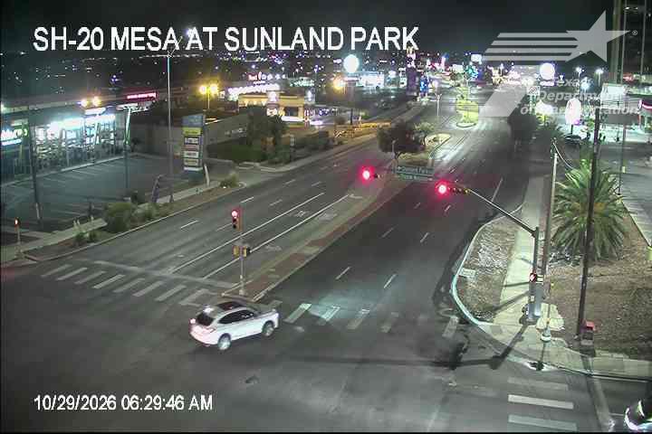 A white SUV is making a left turn at an intersection under red traffic lights, with a wide multi-lane road and commercial buildings visible in the background under nighttime lighting.