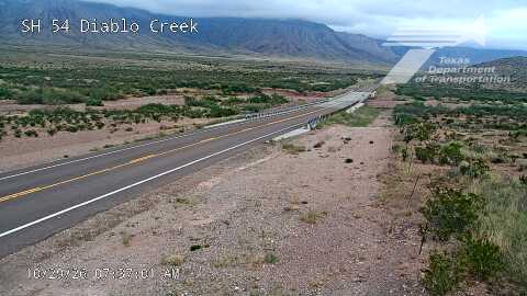 A wide, empty highway stretches through a dry, mountainous landscape with sparse vegetation under an overcast sky.
