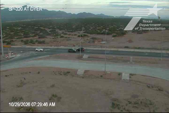 A quiet intersection with light traffic, featuring a white sedan and two dark vehicles near a crosswalk, set against a backdrop of arid terrain and distant mountains under clear skies.
