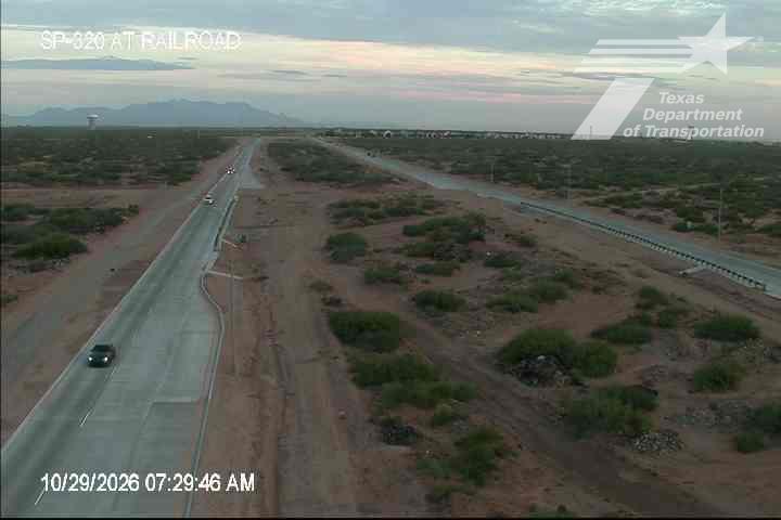 A single dark vehicle travels along a newly paved highway through a desert landscape with sparse vegetation and distant mountains under a soft, overcast sky.