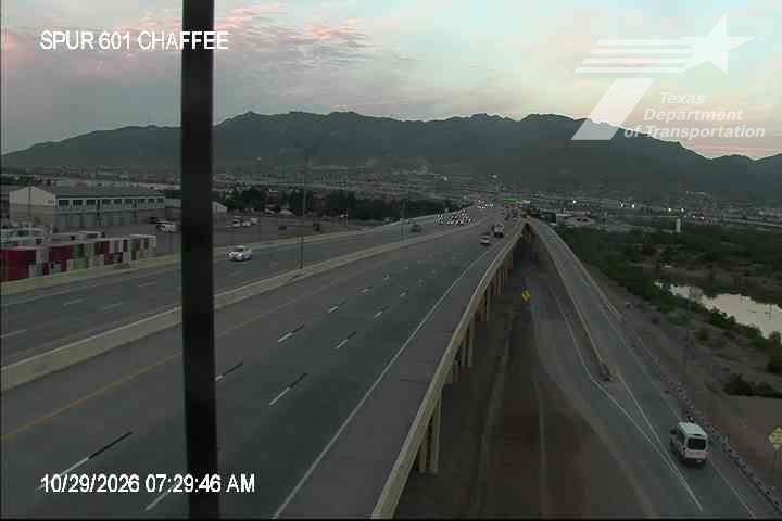 A multi-lane highway with light traffic flows under a clear sky, flanked by distant mountains and a riverbank with minimal construction activity.