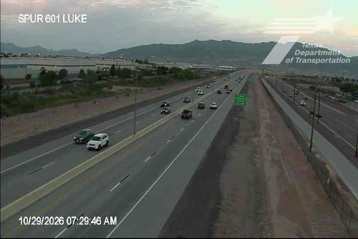 Light traffic flows along a multi-lane highway with distant mountains in the background under clear skies.