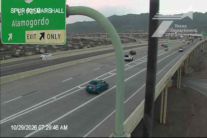 A multi-lane highway with moderate traffic flows under clear skies, featuring a green directional sign for Spur 60, Marshall, and Alamogordo, with distant mountains in the background.