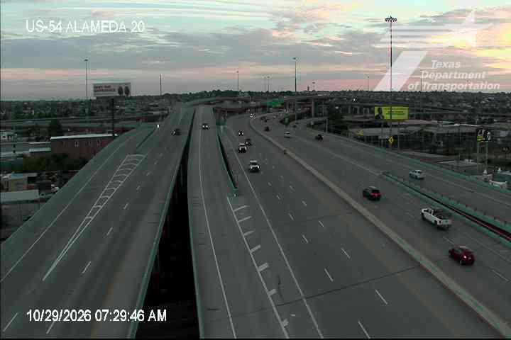 Multi-lane highway with moderate traffic flowing in both directions under a partly cloudy sky, featuring an overpass and distant commercial signage.