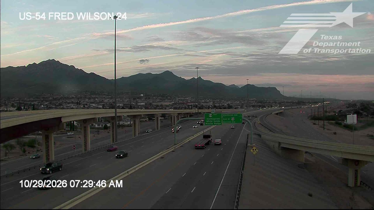 A multi-lane highway with moderate traffic flows under an overpass, with distant mountains visible under a partly cloudy sky at dawn.