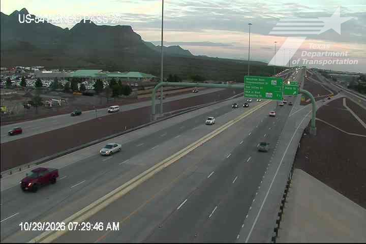 Multi-lane highway with moderate traffic flow under clear skies, bordered by mountains and a commercial area with visible signage.
