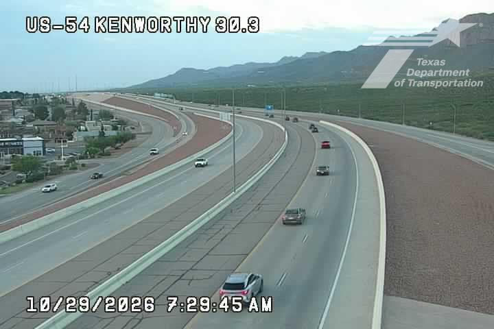 Light traffic flows along a multi-lane highway with distant mountains in the background under clear skies.
