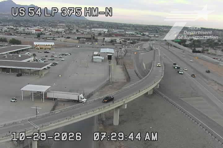 A highway overpass curves over a commercial area with a semi-truck parked at a service station, while light traffic moves along the road under clear skies.