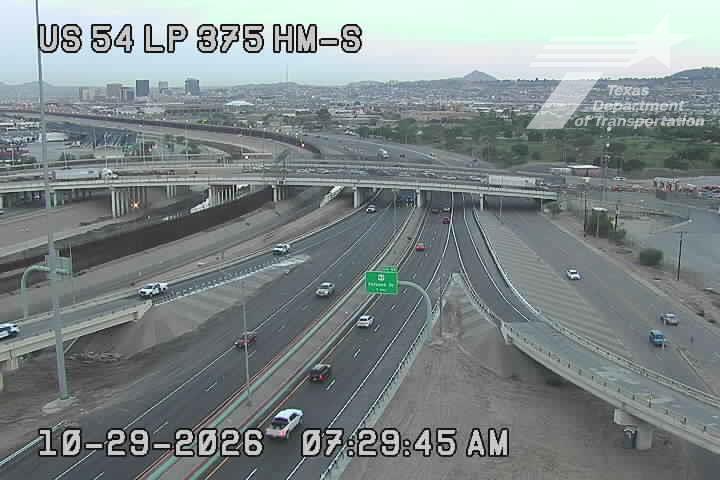 Multi-lane highway with moderate traffic flowing in both directions under an overpass, with no visible incidents or road work; distant city skyline and hazy sky suggest mild weather conditions.