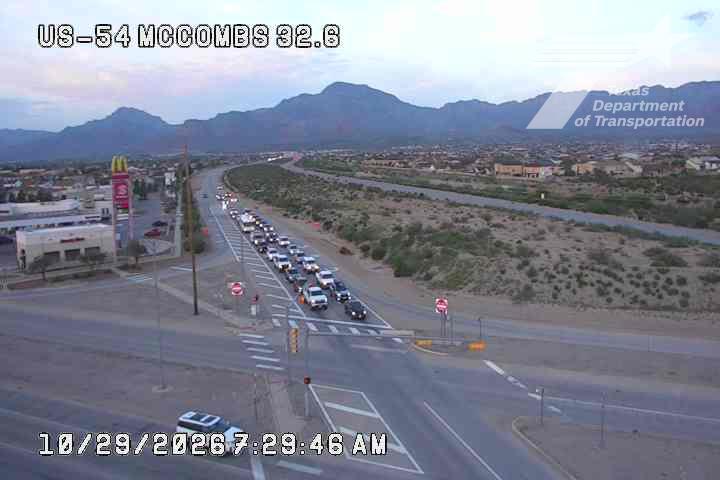 Long line of vehicles moving slowly through an intersection near a McDonald's, with mountains in the background under clear skies.