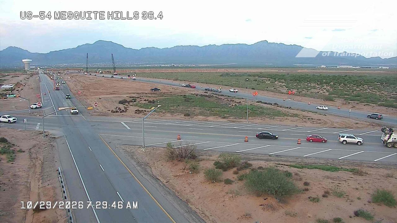 A wide view of a highway intersection with light traffic, surrounded by desert terrain and distant mountains, with road construction activity and traffic cones visible.