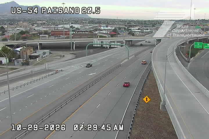 Light traffic flows on a multi-lane highway with a few vehicles visible, including a red car and a black sedan, under clear skies with distant mountains in the background.