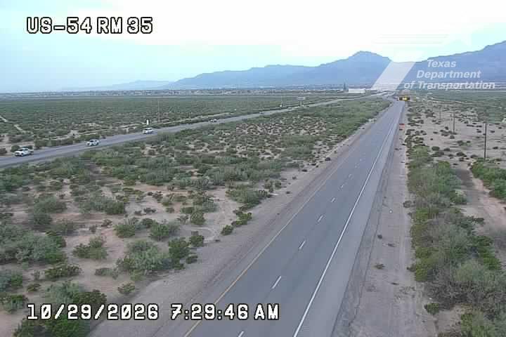 A wide, multi-lane highway stretches through a dry, scrub-covered landscape with distant mountains under clear skies; light traffic moves steadily in both directions with no visible incidents or road work.