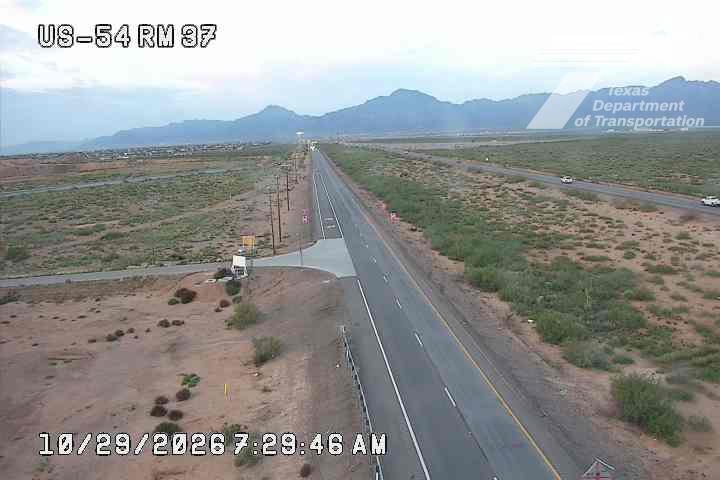 A wide, multi-lane highway stretches through a dry, arid landscape with distant mountains under a cloudy sky; light traffic moves in both directions with no visible incidents or road work.