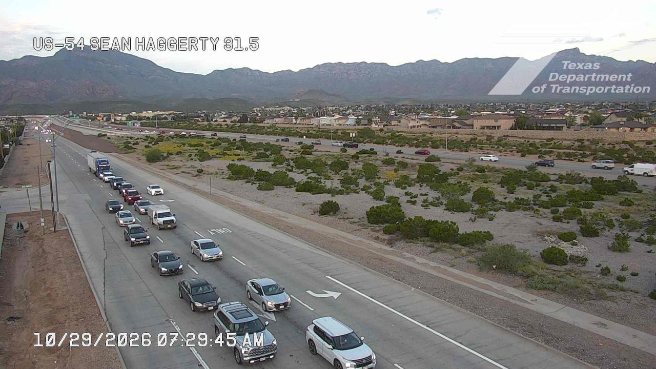 Light traffic flows along a multi-lane highway with vehicles moving at moderate speeds, bordered by desert scrub and distant mountains under clear skies.