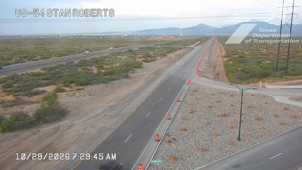 A wide highway stretches through a dry, arid landscape with sparse vegetation, featuring light traffic and construction cones along the right lane, indicating ongoing road work; distant mountains and a water tower are visible under a clear sky.