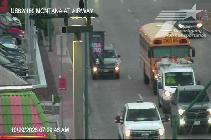 A yellow school bus and multiple vehicles, including a white pickup truck and a van, are moving slowly in traffic on a multi-lane road with a commercial building on the left.