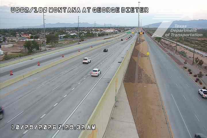 Light traffic flows on a multi-lane highway with construction cones and barriers indicating active road work; distant mountains frame the scene under clear skies.