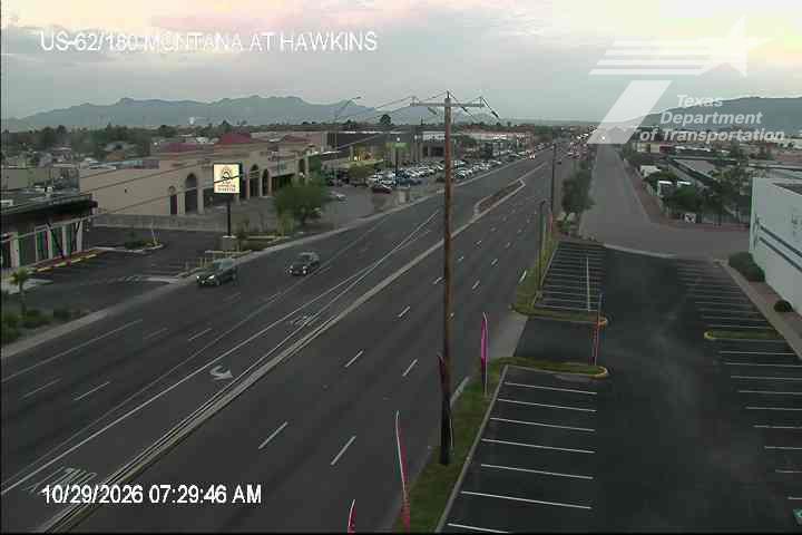 Light traffic flows along a multi-lane highway with commercial buildings on the left and empty parking lots on the right, under a clear sky with distant mountains in the background.