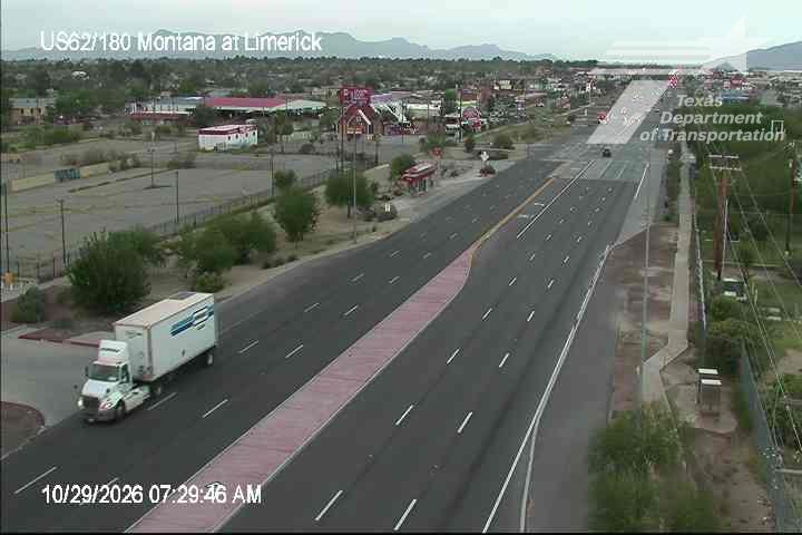 A wide highway with light traffic features a white semi-truck on the left lane, with distant vehicles and commercial buildings along the roadside under an overcast sky.