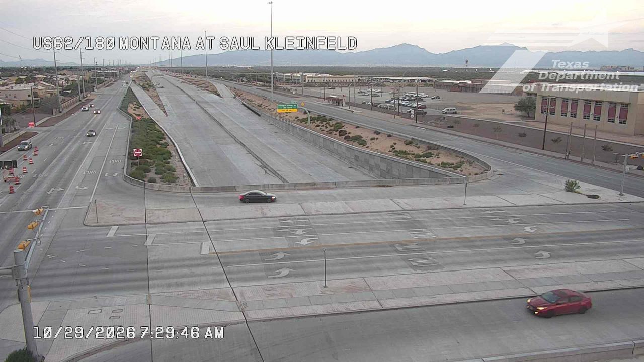 A wide highway intersection with light traffic, featuring a black sedan and a red car in motion, under clear skies with distant mountains visible.