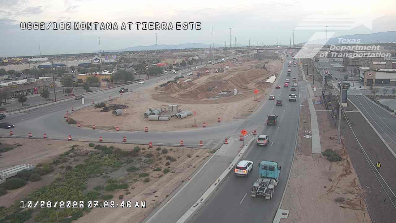 A highway intersection with active road construction, featuring traffic moving slowly through a work zone with cones and barriers; a blue semi-truck and several cars are visible on the road, with a worker in a yellow vest standing near the roadside.