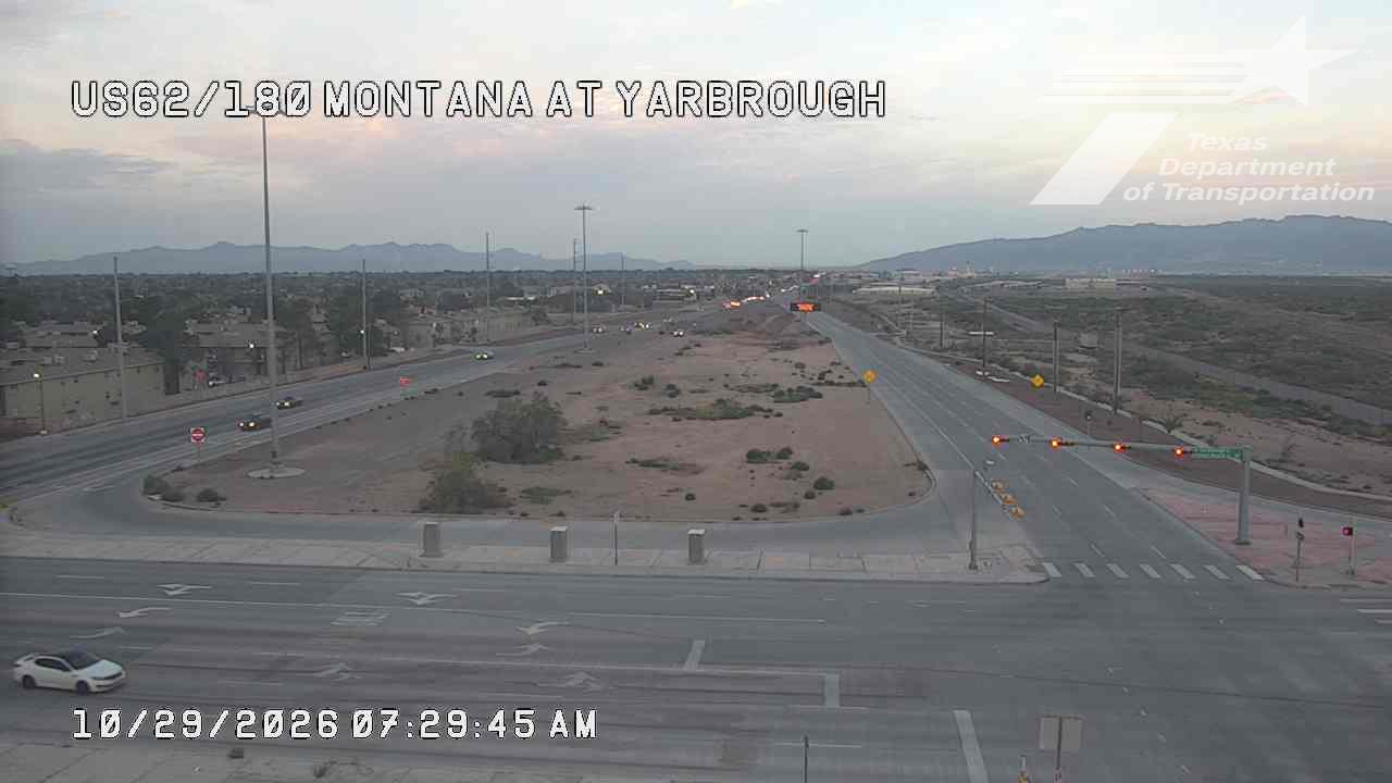 Light traffic flows through a wide intersection at dawn, with a white sedan turning left and distant vehicles moving along the highway; dry, open terrain surrounds the road with low hills in the background.