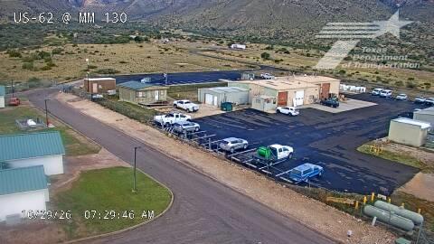 Aerial view of a roadside maintenance facility with multiple parked vehicles and a large paved lot, surrounded by arid terrain and distant mountains under clear skies.