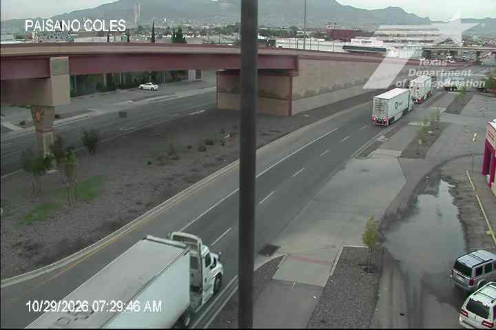A white semi-truck travels under an overpass on a multi-lane highway with light traffic; a line of trucks waits at a traffic signal, and standing water pools near the roadside.