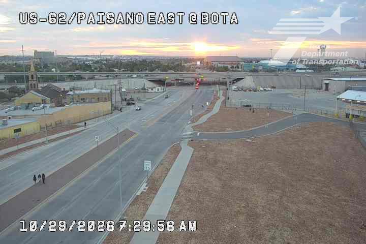 A wide view of a multi-lane highway at dawn with light traffic moving through an intersection under an overpass, and a few pedestrians walking on the sidewalk.