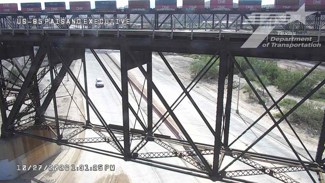 A white vehicle travels on a multi-lane highway beneath a large steel overpass carrying a freight train; the scene is clear and dry with sparse vegetation on the sides.